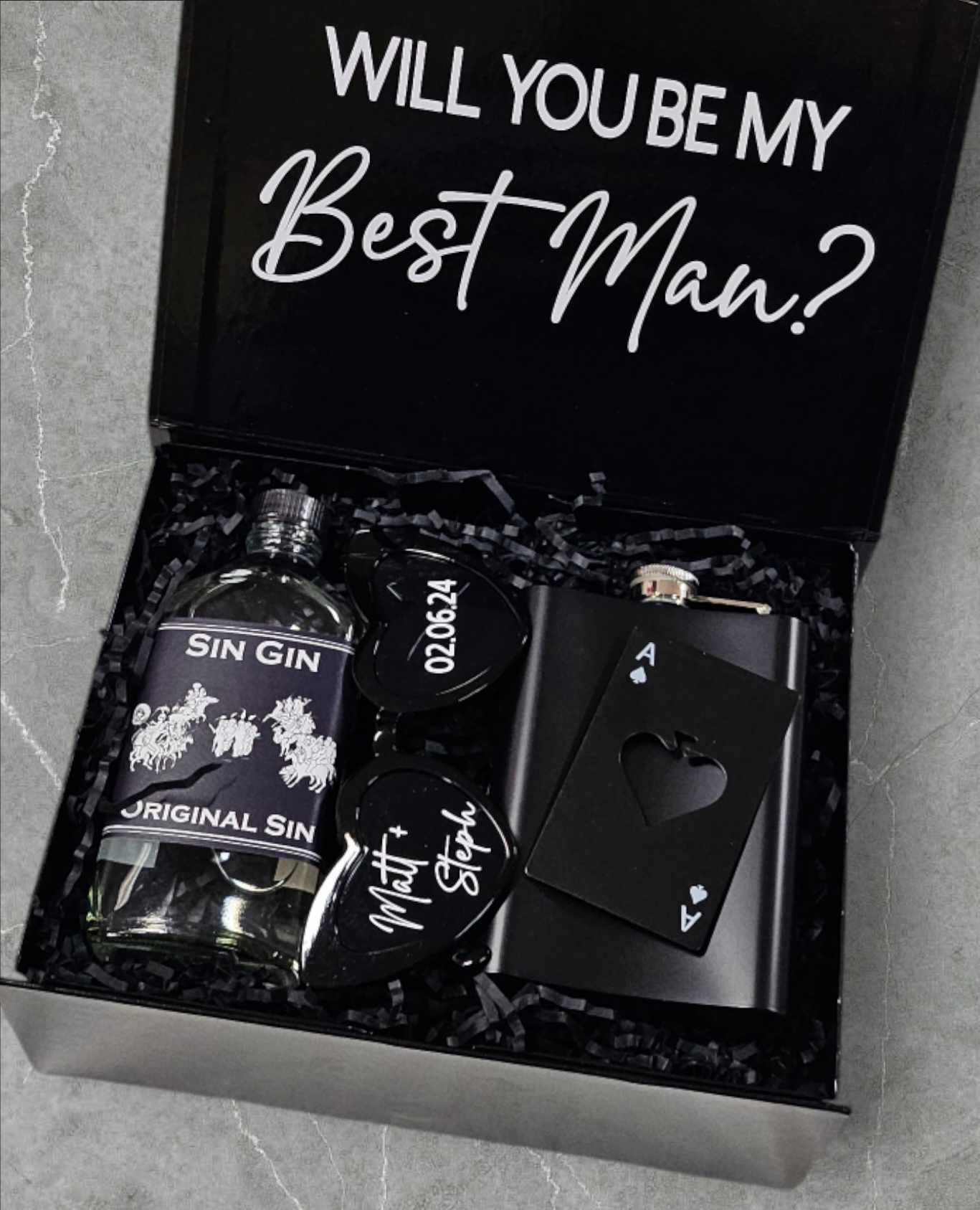 A stylish black magnetic box containing a 200ml bottle of Sin Gin, personalized black heart glasses with a wedding date sticker, a black flask, and a metal playing card bottle opener, all topped with colourful confetti.