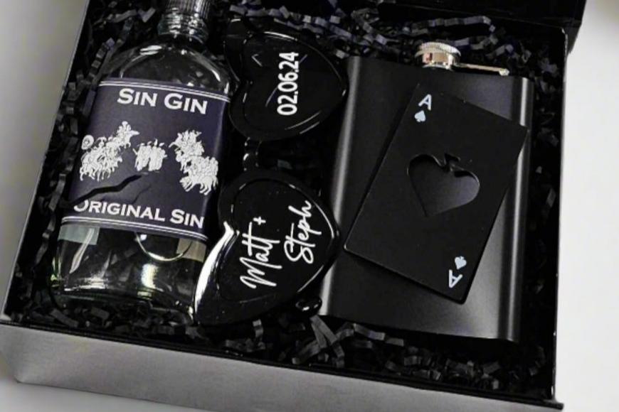 A stylish black magnetic box containing a 200ml bottle of Sin Gin, personalized black heart glasses with a wedding date sticker, a black flask, and a metal playing card bottle opener, all topped with colorful confetti.