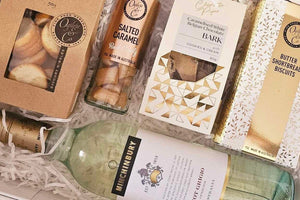 Nibble & Sip Delight Hamper: A luxurious selection of savory and sweet treats, including Ogilvie & Co Garlic Nibbles, Salted Caramel Rock Candy, Charlotte Pipe Caramelised White Belgian Chocolate Bark, Ogilvie Shortbread Biscuits, and Michinbury Pinot Grigio. Packaged in a white magnetic close box, topped with signature confetti and a bow for a chic and indulgent experience.