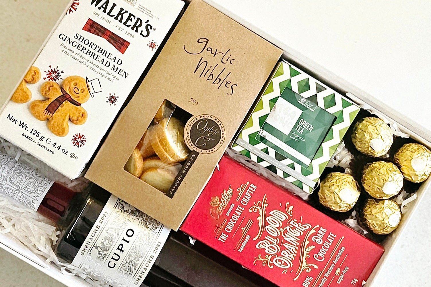 A Christmas hamper featuring Cupio Pinot Noir wine, Walkers shortbread biscuits, Ogilvie garlic nibbles, 5 Ferrero Rocher chocolates, Crunch Box blood orange chocolate, and a box of green tea, all beautifully packaged in a white magnet gift box.