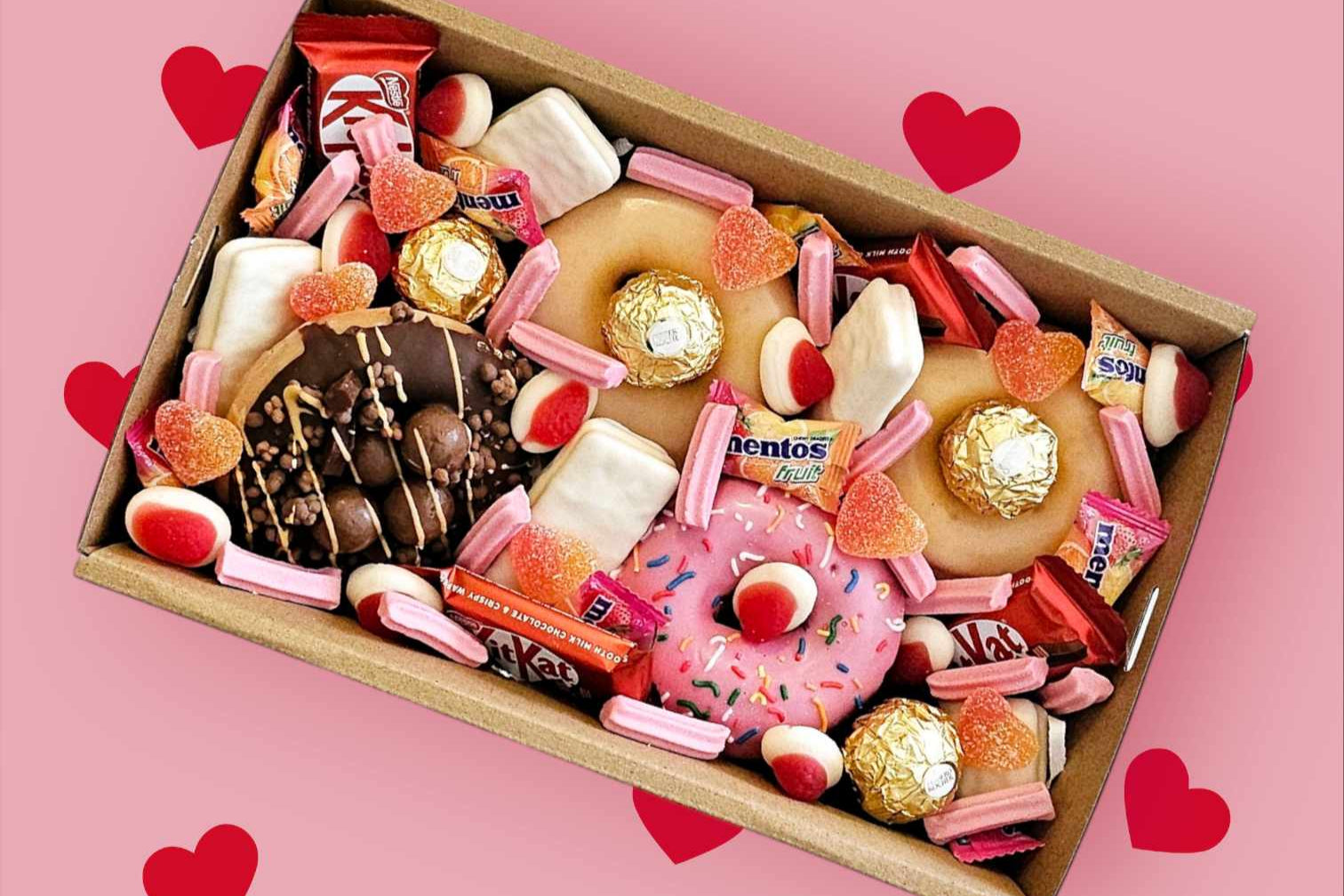 Valentine’s Sweet Treat Box featuring 4 Krispy Kreme donuts topped with KitKat Minis, white TimTams, sour heart lollies, mini musk sticks, strawberries & cream Allens lollies, Mentos, and Ferrero Rocher chocolates, presented in a 25x16x8 cm gift box.