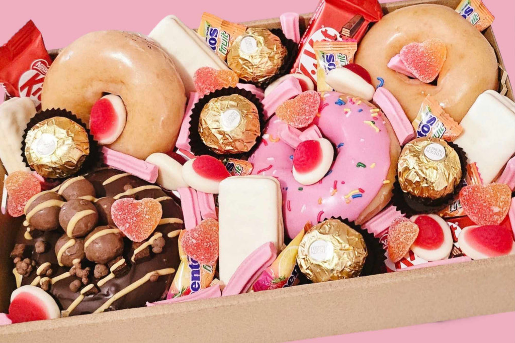 Valentine’s Sweet Treat Box featuring 4 Krispy Kreme donuts topped with KitKat Minis, white TimTams, sour heart lollies, mini musk sticks, strawberries & cream Allens lollies, Mentos, and Ferrero Rocher chocolates, presented in a 25x16x8 cm gift box.