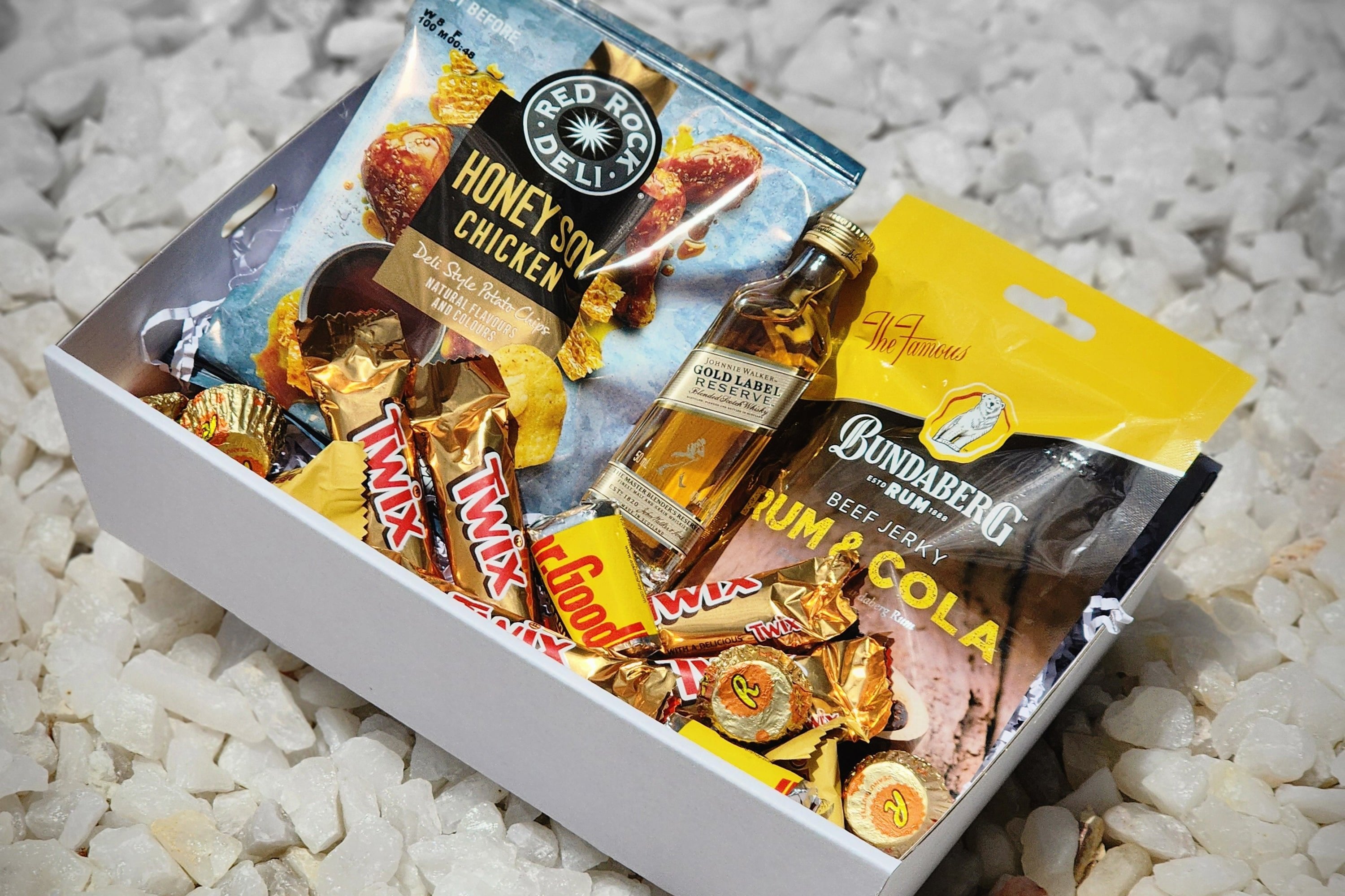 A premium Gold Label gift box featuring a 50ml bottle of Johnnie Walker Gold Label, Red Rock Deli Honey Soy chips, Dr. Proctor's Bundy Rum & Cola beef jerky (30g), five mini Twix bars, four mini Hershey's chocolates, four Reese's Peanut Butter Cups, and two mini Toblerone chocolates, all elegantly arranged for a sophisticated treat.